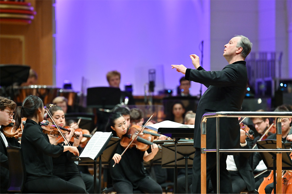 A man, wearing formal attire, conducting an orchestra with his hands, with students performing on their instruments, also wearing formal attire.
