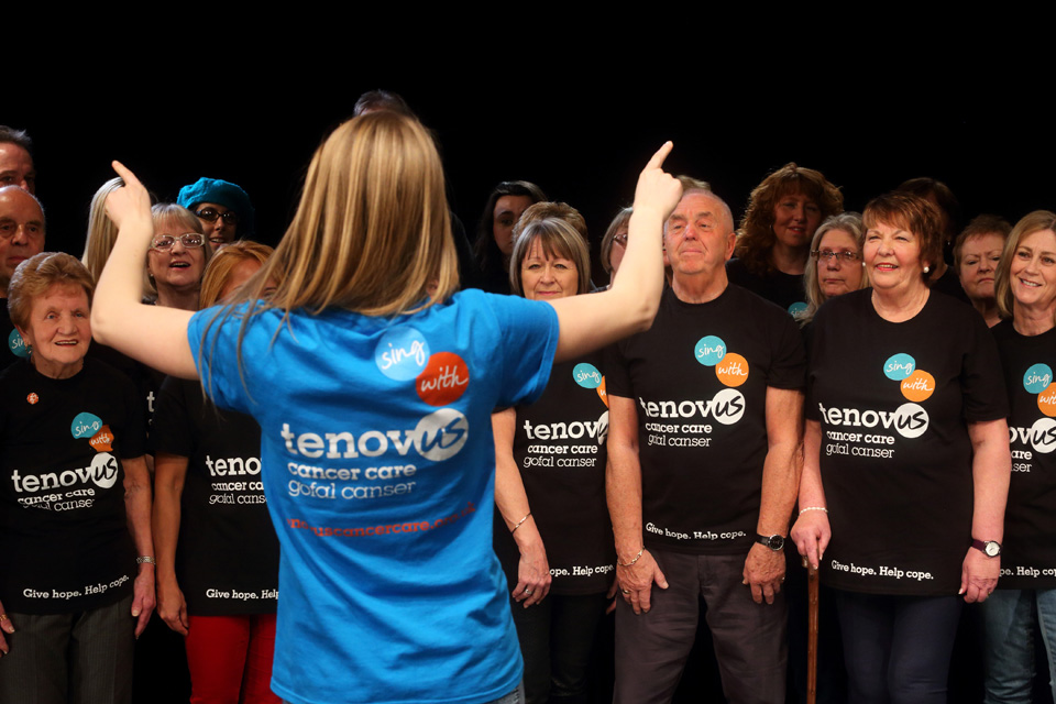 A group of people singing, with a lady with blonde hair, conducting the group of singers.
