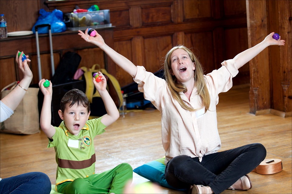 A white women, singing and playing the maracas in a group, with a young boy sitting next to her.