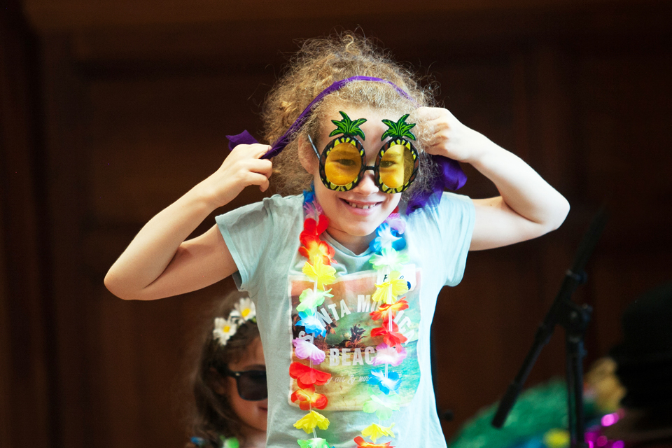 A young girl with light curly hair, wearing glasses shaped like pineapples, smiling at the camera.