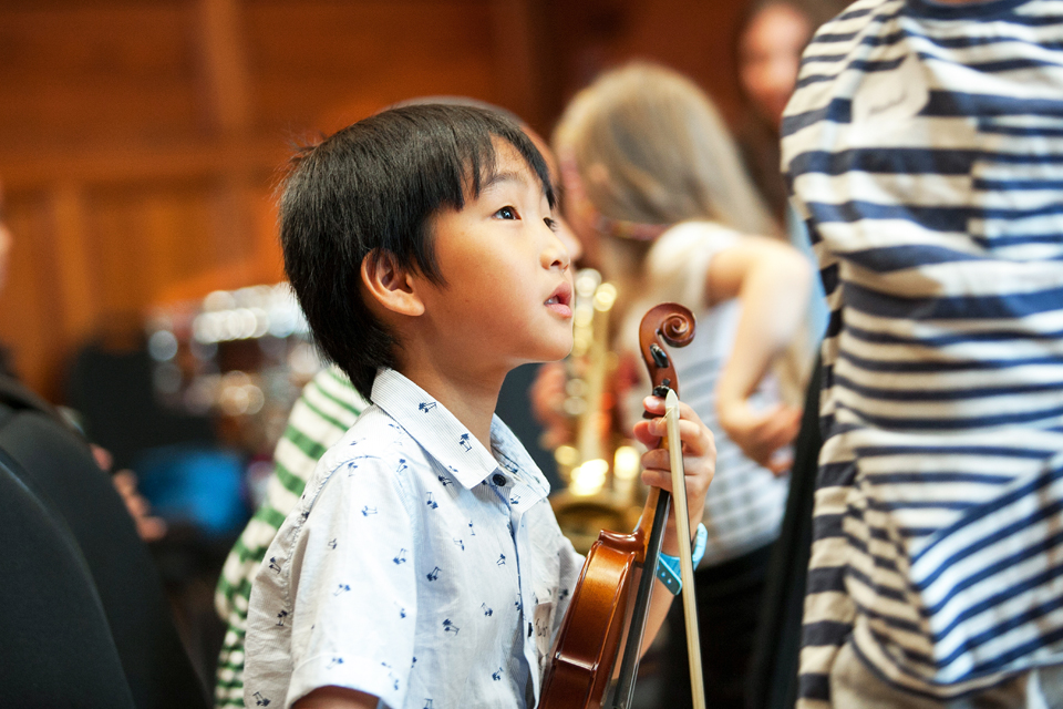 An Asian boy, wearing a light blue shirt, holding a violin, in his hands.