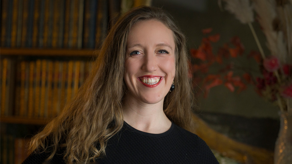 A women with long hair, wearing black smart attire, smiling at the camera, with a wood-panelled background behind her. A women with long hair, wearing black smart attire, smiling at the camera, with a wood-panelled background behind her.