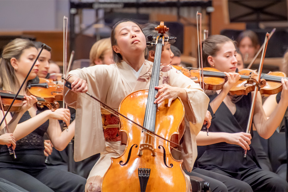 An Asian female student, wearing a formal kimono, playing the cello, with students performing behind her.