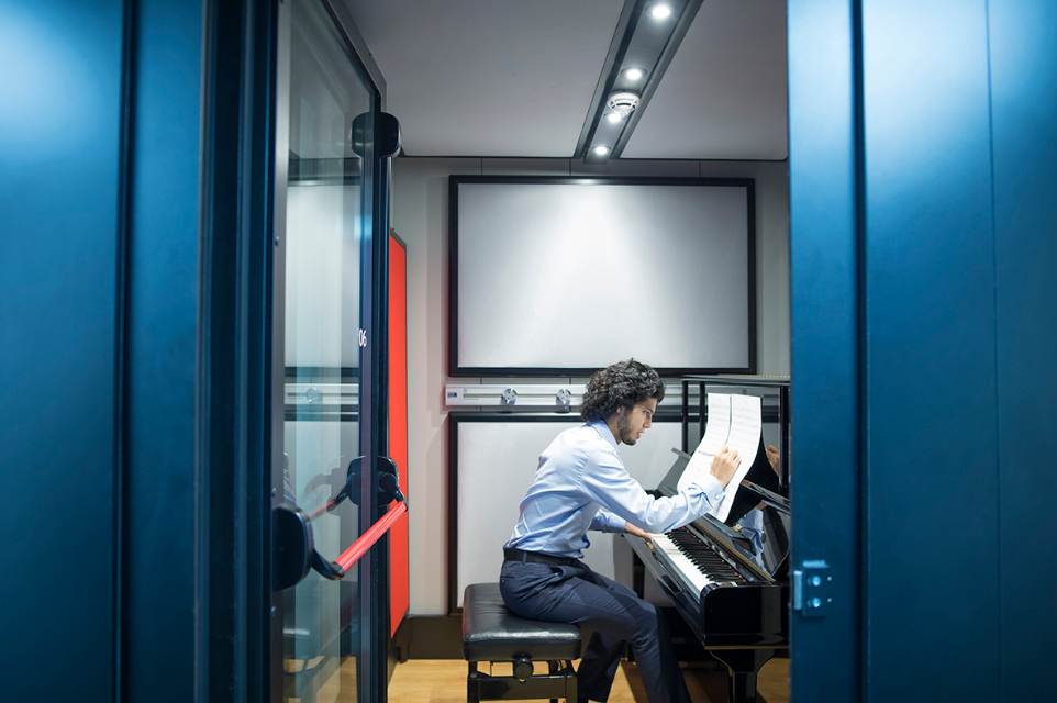 A mixed race male student, sitting on a piano stool, make a note on some music sheets, propped up on the black piano, sitting in an open practice pod.