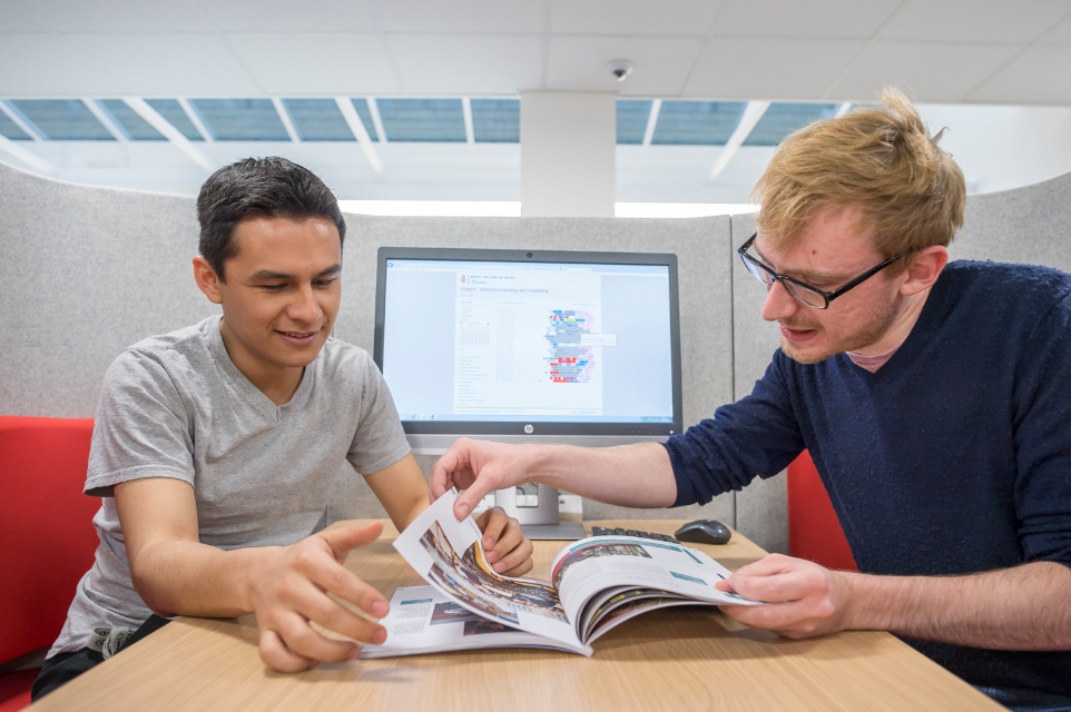 Two male students, one with glasses, looking through a prospectus, sitting in a group study pod.