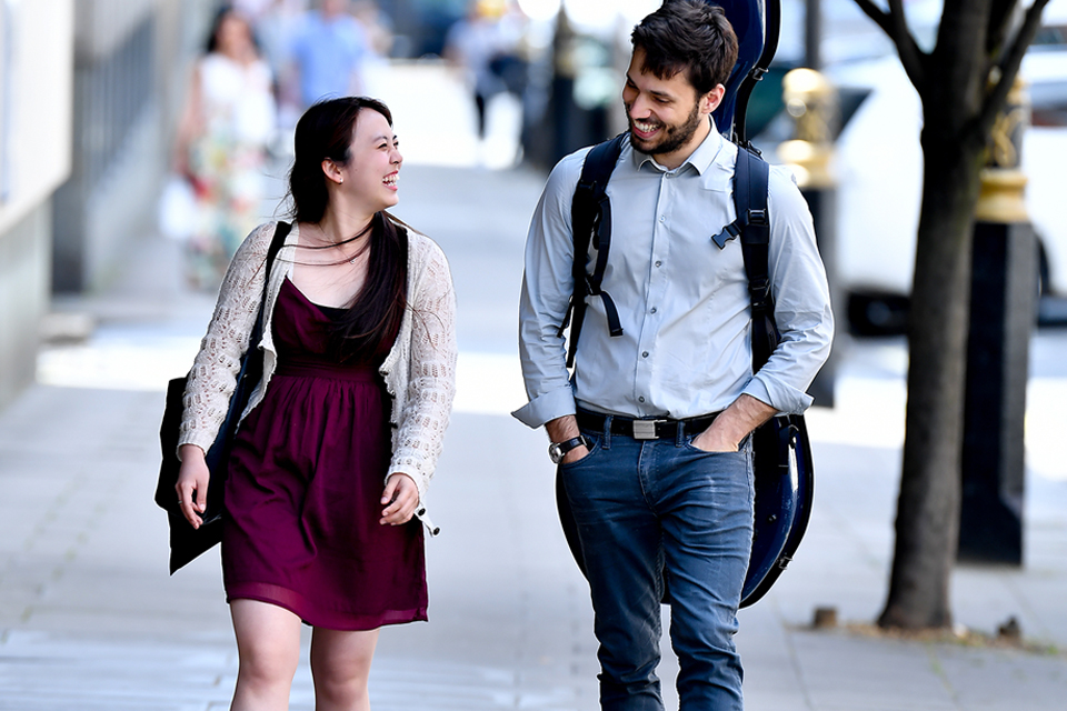 Two students, carrying a bag and music instrument, smiling and chatting, walking towards the college.