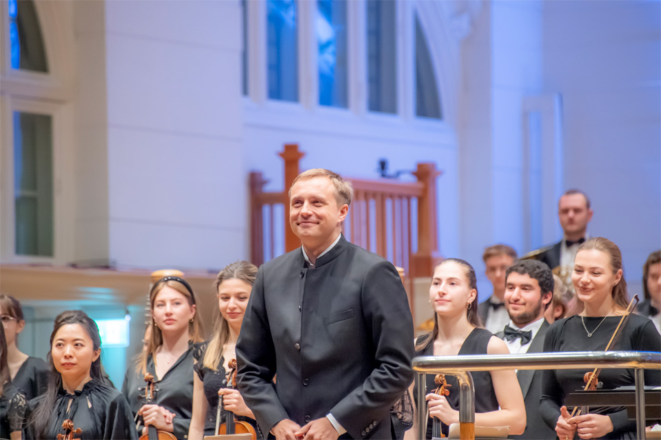 A man, wearing formal attire, smiling and facing the audience, standing outside the conductor's podium, with students standing next to and behind him, smiling and holding their instruments.