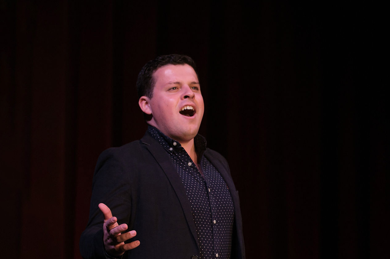 A male student, wearing a dark patterned shirt and a blazer, singing on stage with red curtains behind him