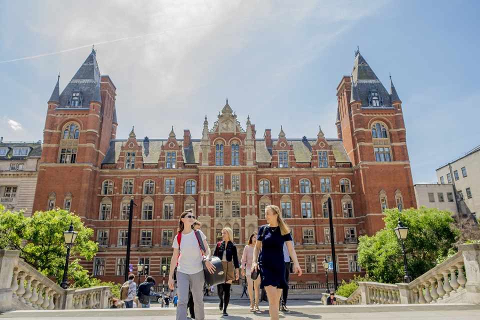 A group of students, smiling and chatting, with the front entrance of the Royal College of Music, an old, red brick, Victorian building, in the background.