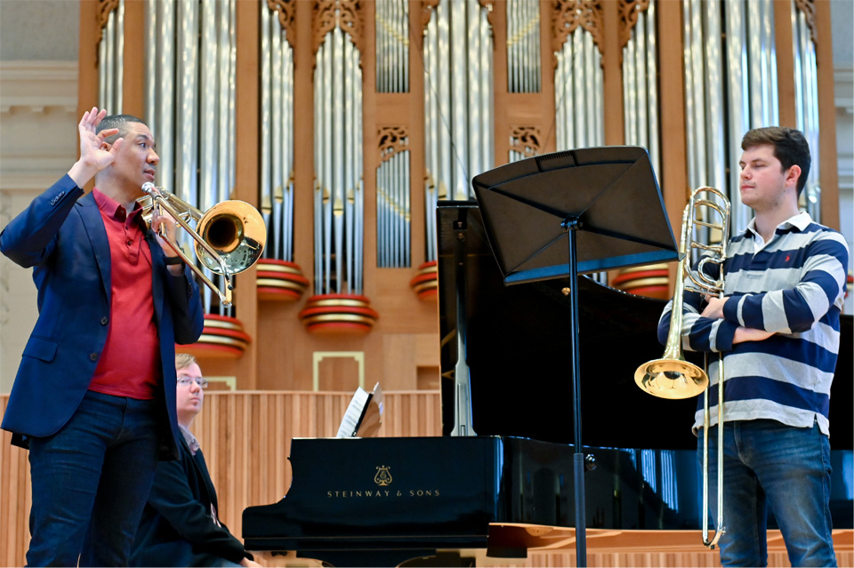 Male student, holding his trombone, standing and participating in a trombone masterclass with Weston Sprott, a black man, holding his trombone and talking to the student.